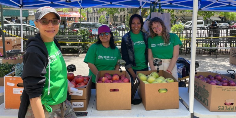 4 people in green shirts standing by a table holding boxes of apples and onions