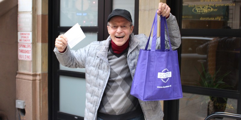 A man in a baseball cap smiling and holding a purple reusable shopping bag that says DOROT.