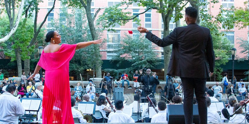 Two people standing on an outdoor stage singing to each other with an orchestra in front of them. Their backs are to the camera.