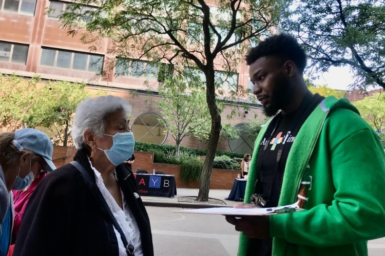 A young man in a green hoodie and an Arts & Minds t-shirt talks to an older woman wearing a mask.