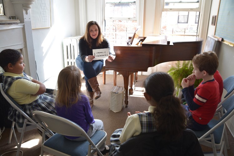Lessons at Bloomingdale School of Music. A woman sitting at a piano shows children a piece of paper with musical notes printed on it.
