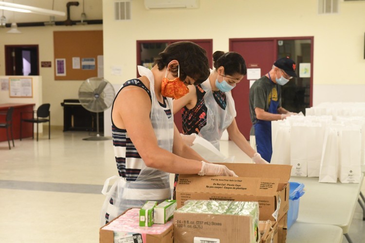 A man in a mask and gloves takes food out of a box. In the background are bags for bagged lunches.