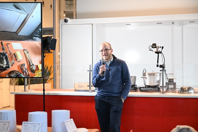 A man wearing glasses leans against a counter with cooking implements on it and speaks into a microphone.