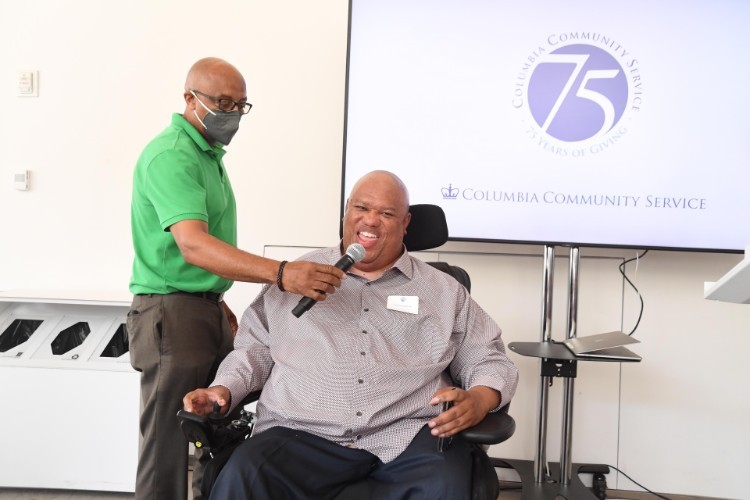 A man in a green shirt holds a microphone for a man in a wheelchair.