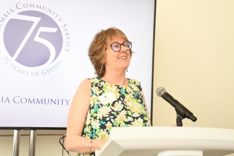 A woman in a floral dress with short hair and glasses stands at a lectern with a microphone