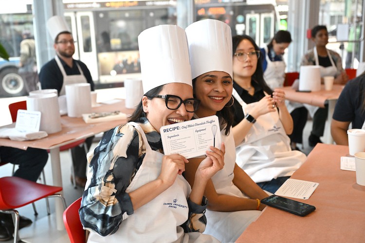 Two women in aprons and chef hats smile at the camera. One is holding up a recipe card.