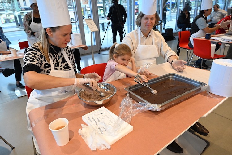 Two women in hats and aprons watch as a child uses an ice cream scoop to scoop up chocolate from a tray.