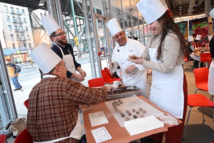 A man in a chef hat and jacket helps three people in aprons and chef hats make truffles.