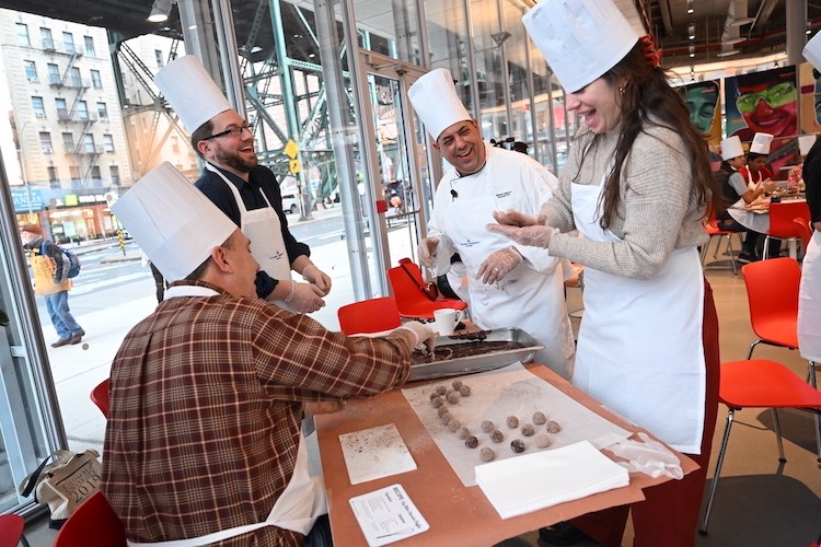 Chef Mike had plenty of help for participants. Photo by Diane Bondareff. A man in a chef hat and jacket helps three people in aprons and chef hats make truffles.