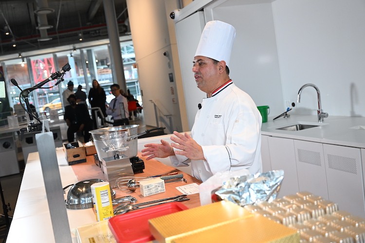 A man in a chef hat and coat speaks while standing behind a counter with cooking supplies and tools on it.