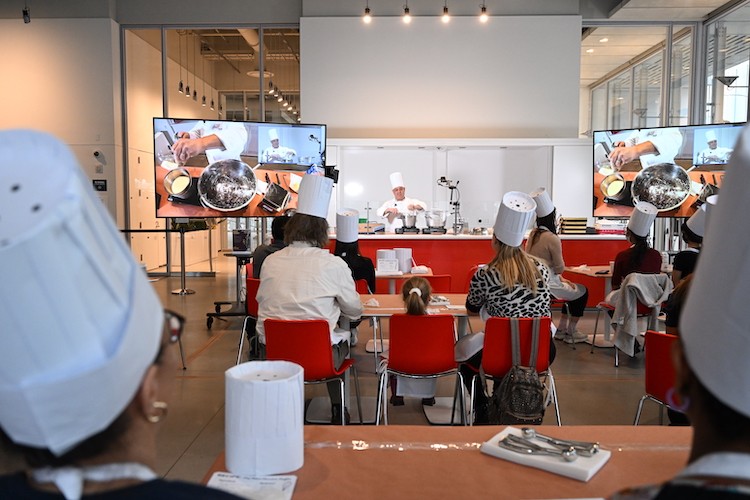 Participants in the event watch Chef Mike, and a close-up of his work bench is visible on screens.