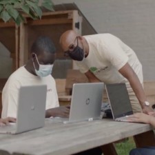 A man in a mask leans over another man in a mask using a laptop computer.