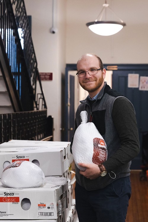 A man holds a packaged turkey, standing by a pile of boxes of turkeys.