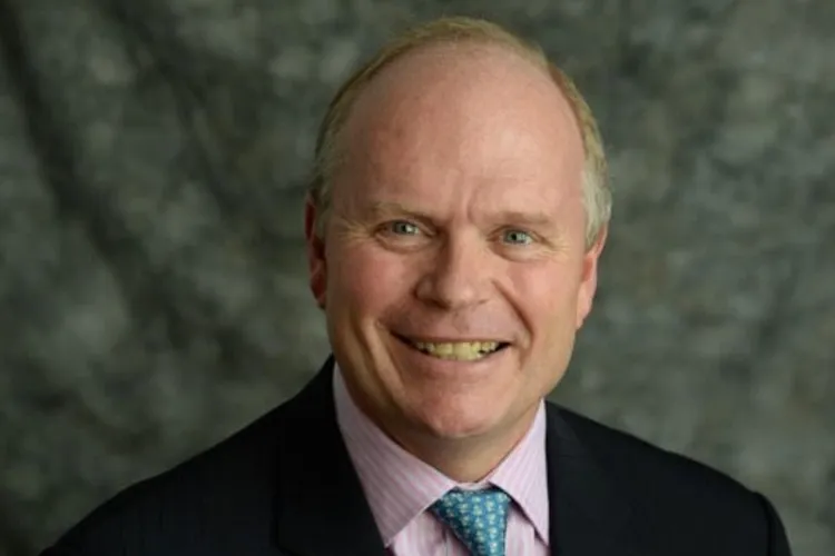 A headshot of a blond man in a pink shirt and black jacket smiling at the camera.