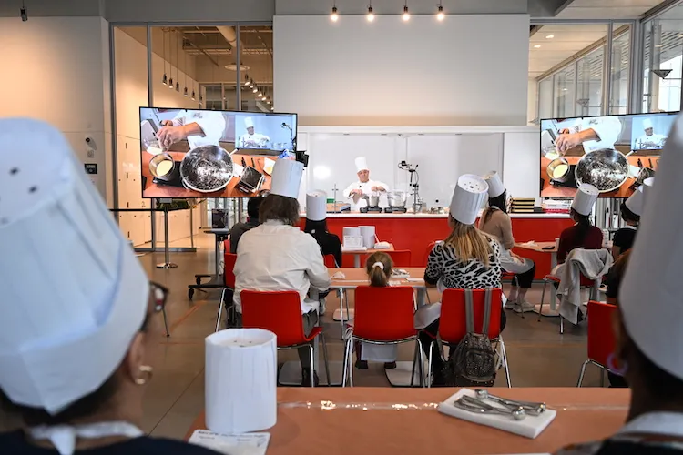 Participants in the event watch Chef Mike, and a close-up of his work bench is visible on screens.